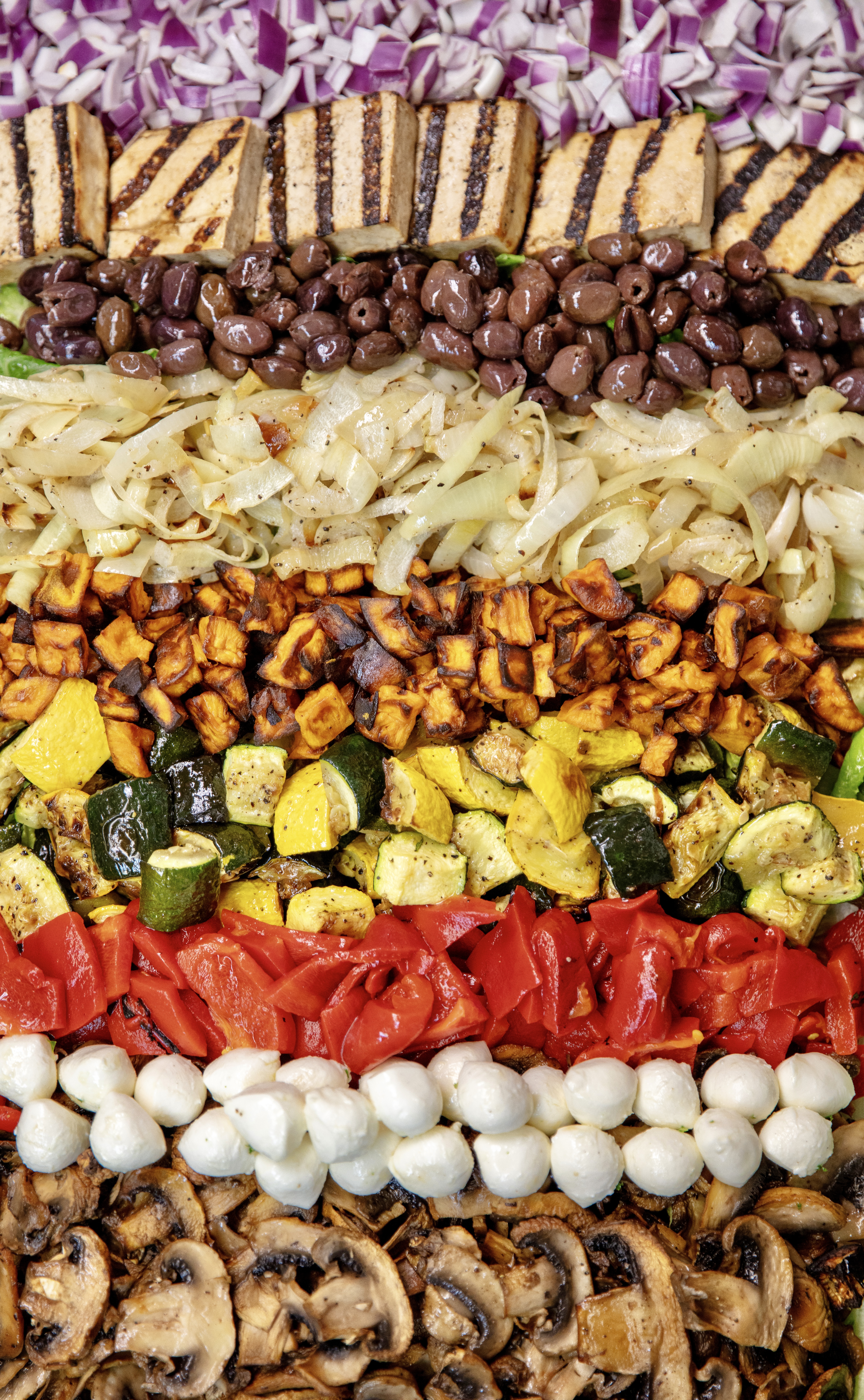 Salad bar toppings including grilled tofu, kalamata olives, roasted onions and vegetables